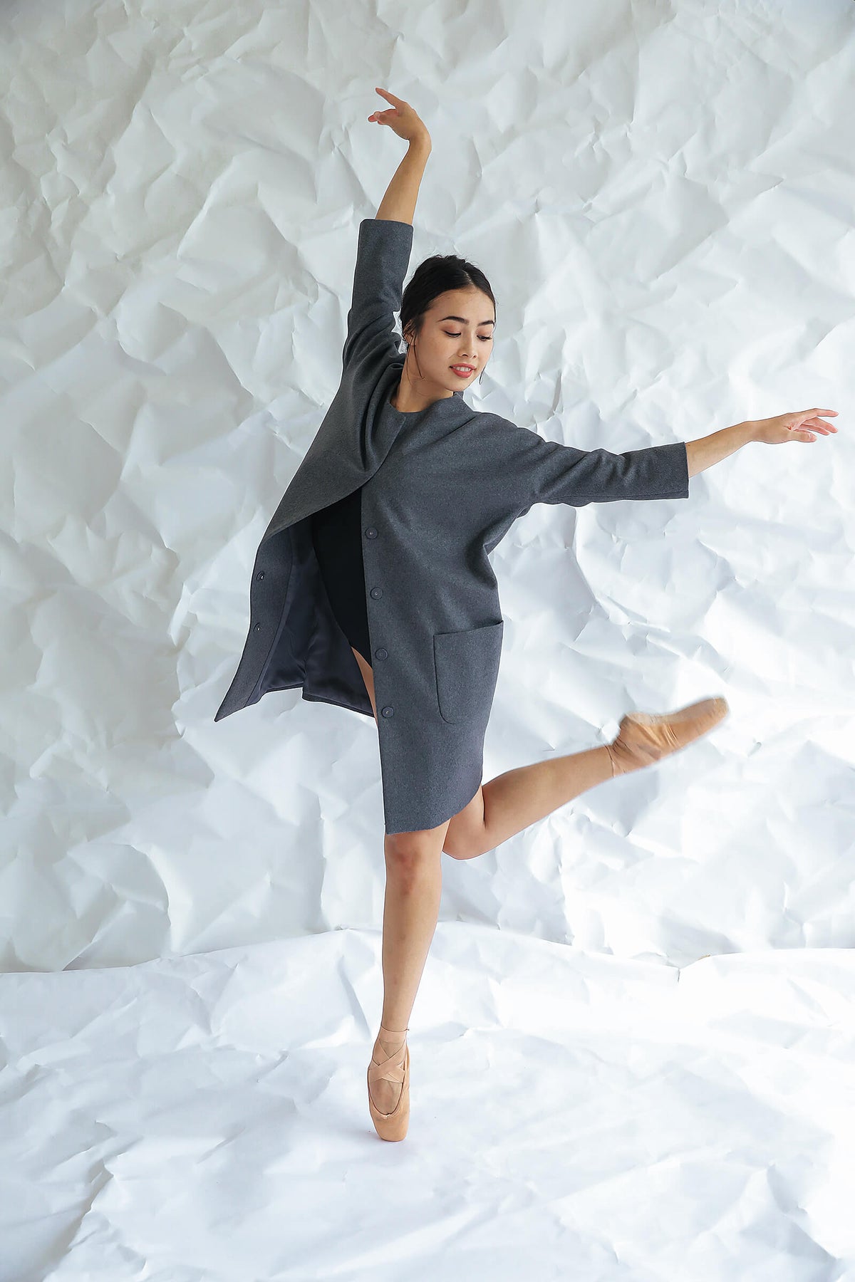 A woman wearing the Battersea grey cashmere coat with raglan sleeves and ballet shoes poses on pointe against a crumpled white paper backdrop, her arms gracefully extended as she balances on one foot.