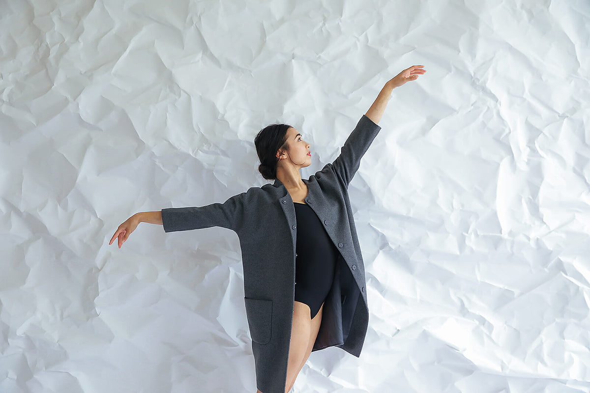 A woman models the Battersea—a grey cashmere coat with raglan sleeves—over a black leotard, posing gracefully with raised arms against a crumpled white paper backdrop.