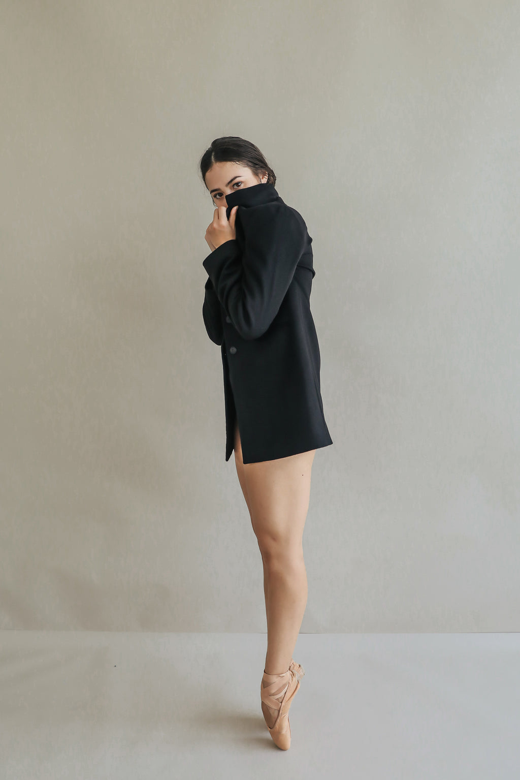 A woman in ballet shoes stands on pointe, wearing Devon, a long black jacket with handmade snap buttons that cover her upper body. Her face is partially obscured by the riding coat as she gazes at the camera against a plain light gray backdrop.