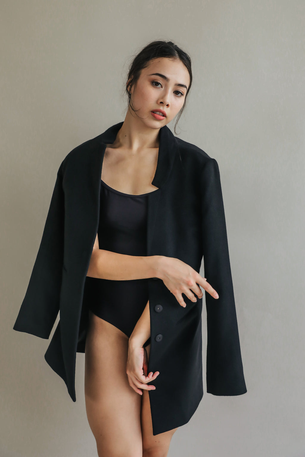 A woman poses against a neutral background, wearing the Devon, a black cashmere coat draped over her shoulders and a black bodysuit. She grips the edge of the coat with one hand to showcase its handmade snap buttons, while the other hand rests on her hip as she looks directly at the camera.