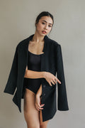 A woman poses against a neutral background, wearing the Devon, a black cashmere coat draped over her shoulders and a black bodysuit. She grips the edge of the coat with one hand to showcase its handmade snap buttons, while the other hand rests on her hip as she looks directly at the camera.