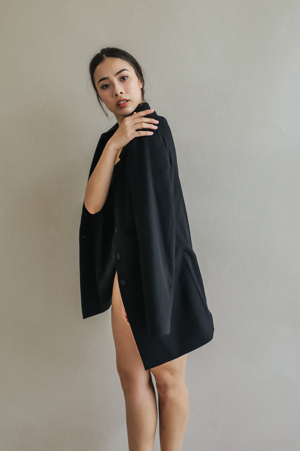 A woman stands against a plain backdrop, draped elegantly in the Devon oversized black blazer with handmade snap buttons. She looks towards the camera with a serious expression, her hair tied back and makeup minimal.