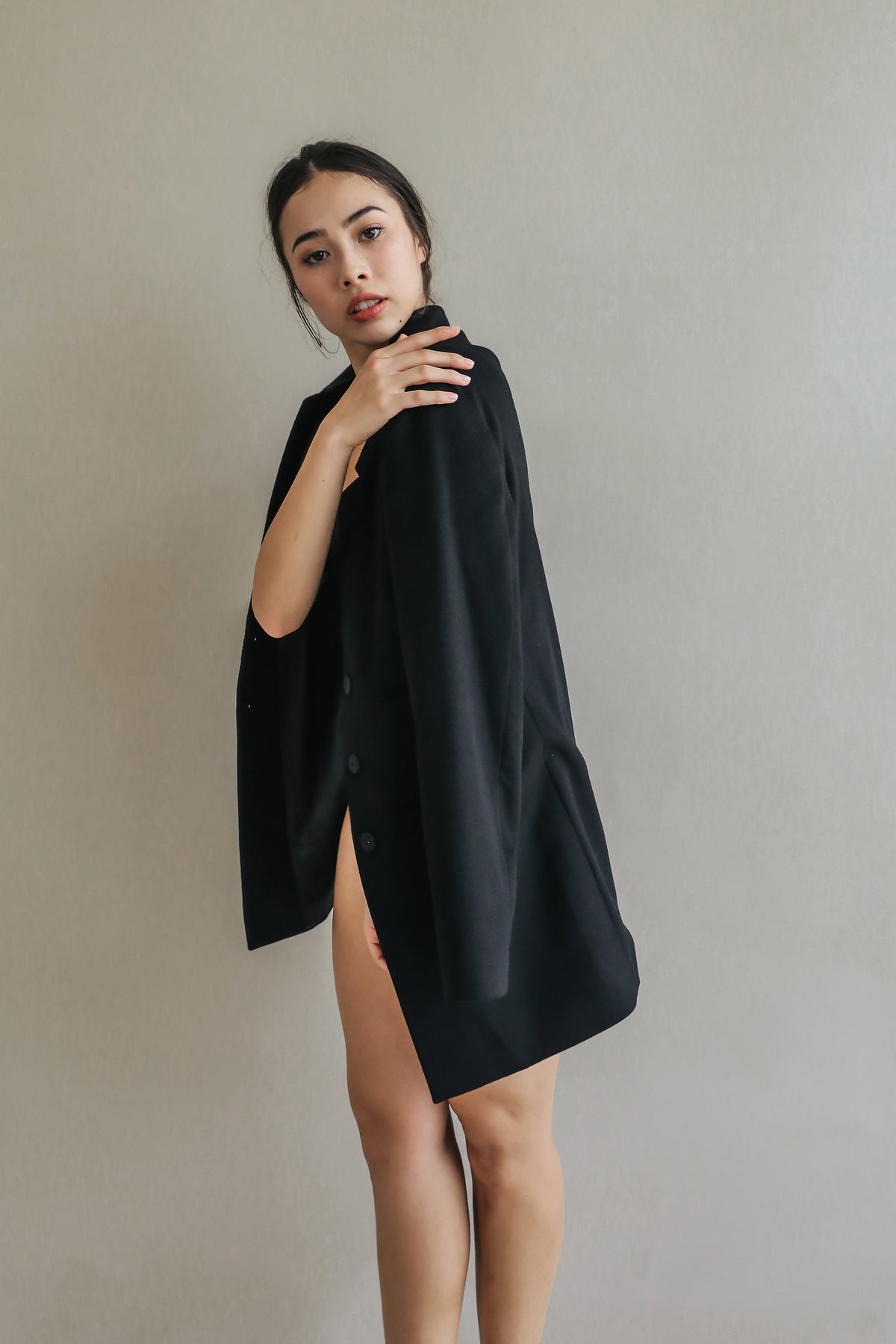 A woman stands against a plain backdrop, draped elegantly in the Devon oversized black blazer with handmade snap buttons. She looks towards the camera with a serious expression, her hair tied back and makeup minimal.