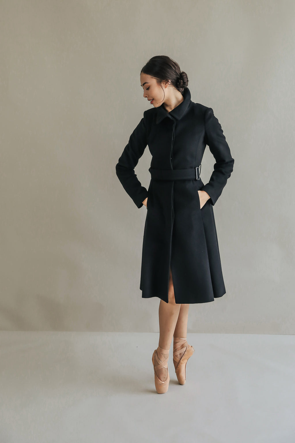 A woman in a Hyde skirted trench coat poses gracefully on pointe shoes against a neutral background. She looks to her left with her hands in the coat pockets, exuding elegance and poise.