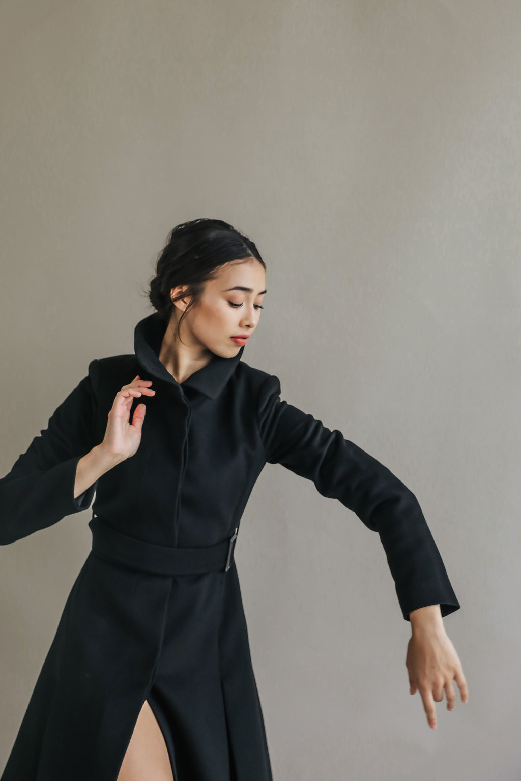 A person in a Hyde black, woven cashmere coat gracefully poses against a plain, light-colored background. They have dark hair pulled back and an elegant expression, with one arm raised and the other lowered, conveying a sense of movement.