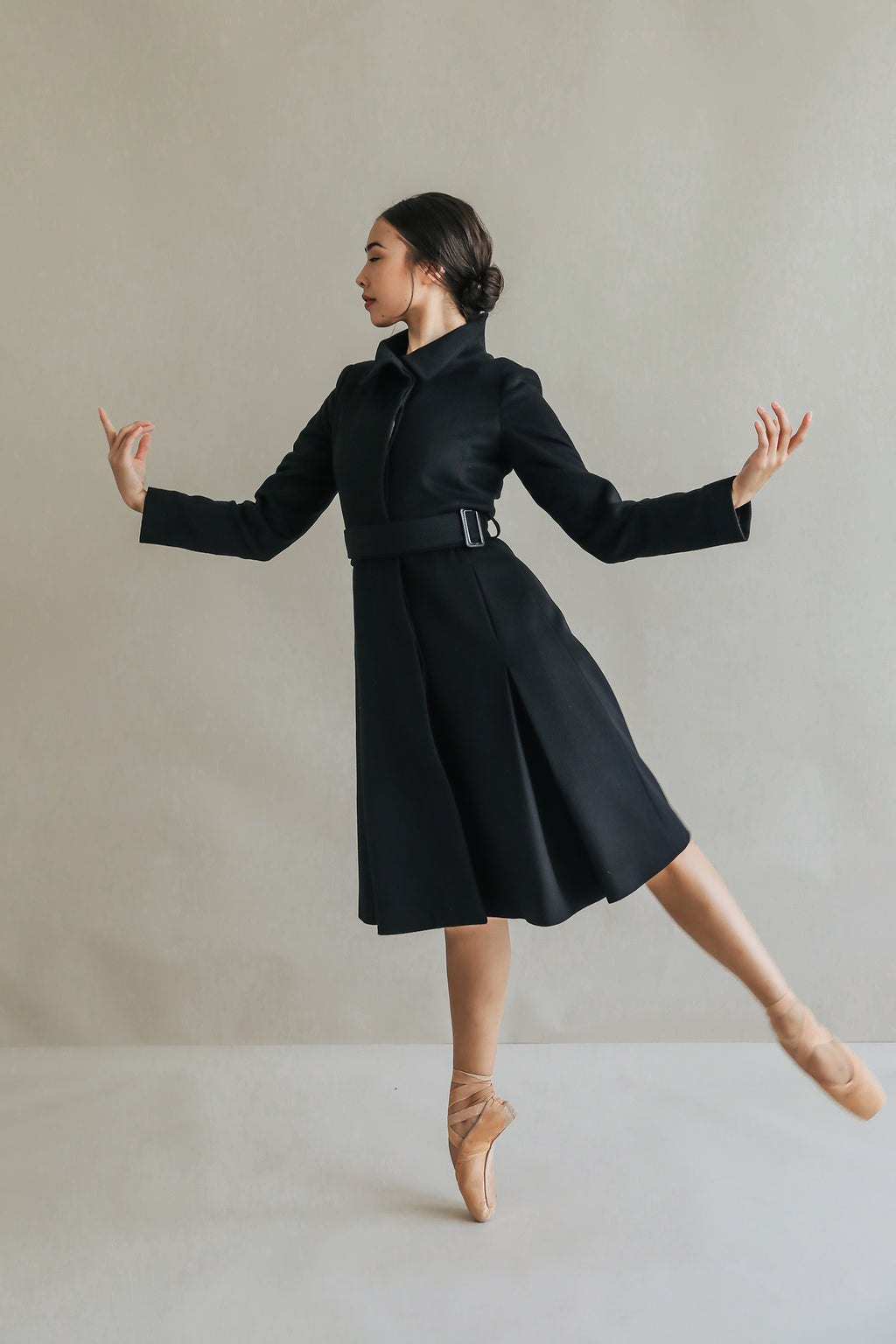 A ballerina in the Hyde woven cashmere skirted trench gracefully balances on pointe, arms extended. The high collar frames her neck as she stands against a neutral background, capturing a moment of elegant poise.