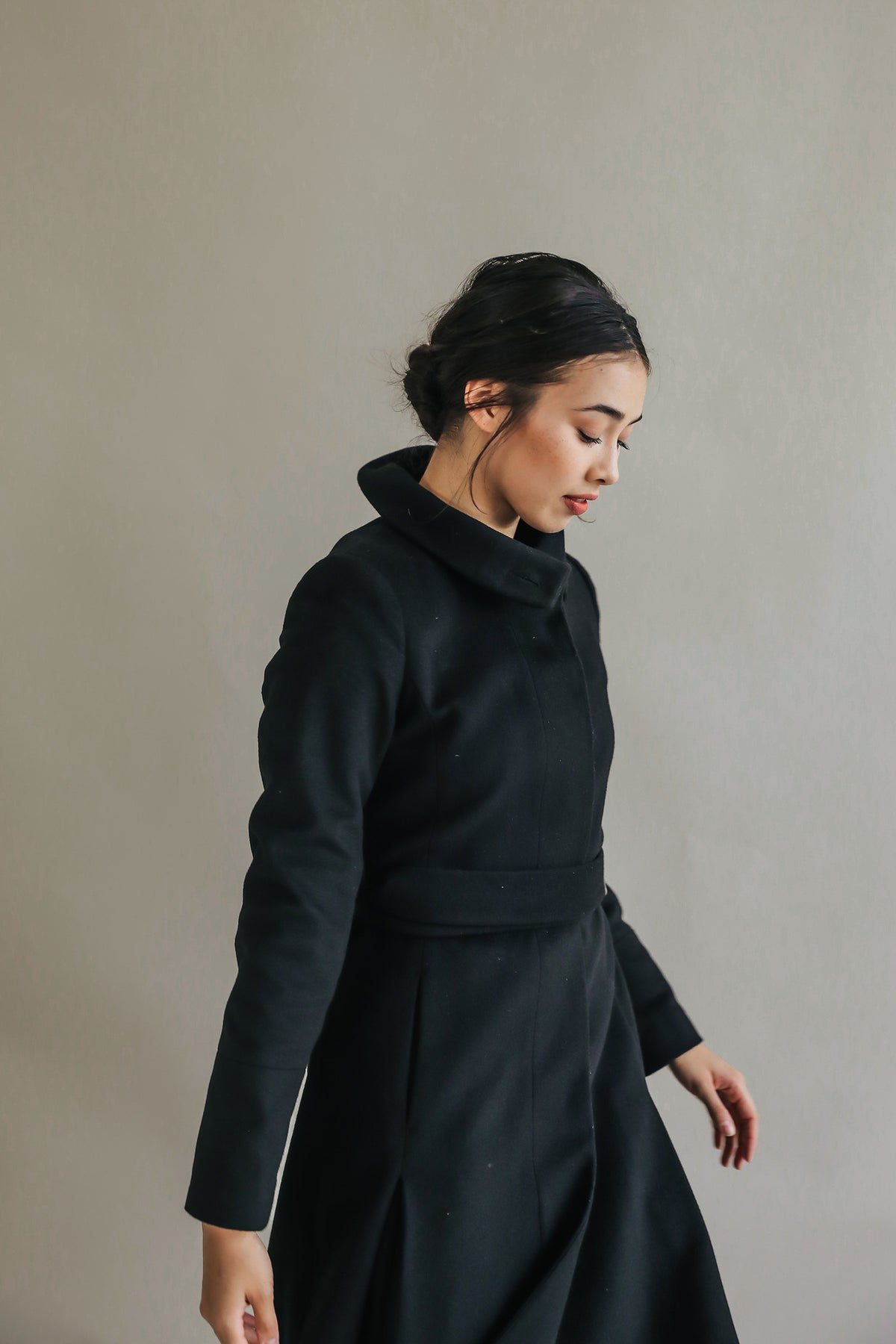 A person with dark hair styled in a bun is wearing the Hyde, a long black skirted trench. They stand against a neutral background, looking down and appearing to be in motion with their hands slightly raised and the high collar subtly framing their face.