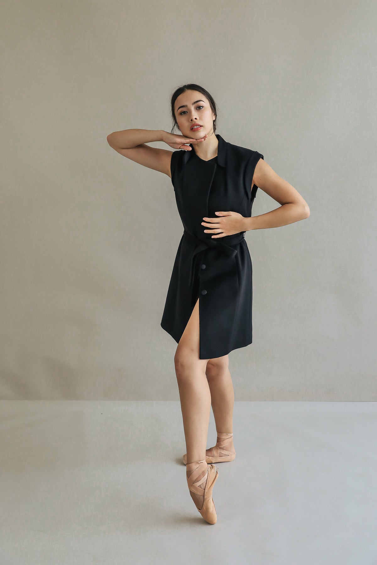 A woman poses gracefully in the Lexington sleeveless black dress with a belted waistline and ballet shoes, standing on pointe against a plain light background, one arm bent under her chin and the other at her waist.