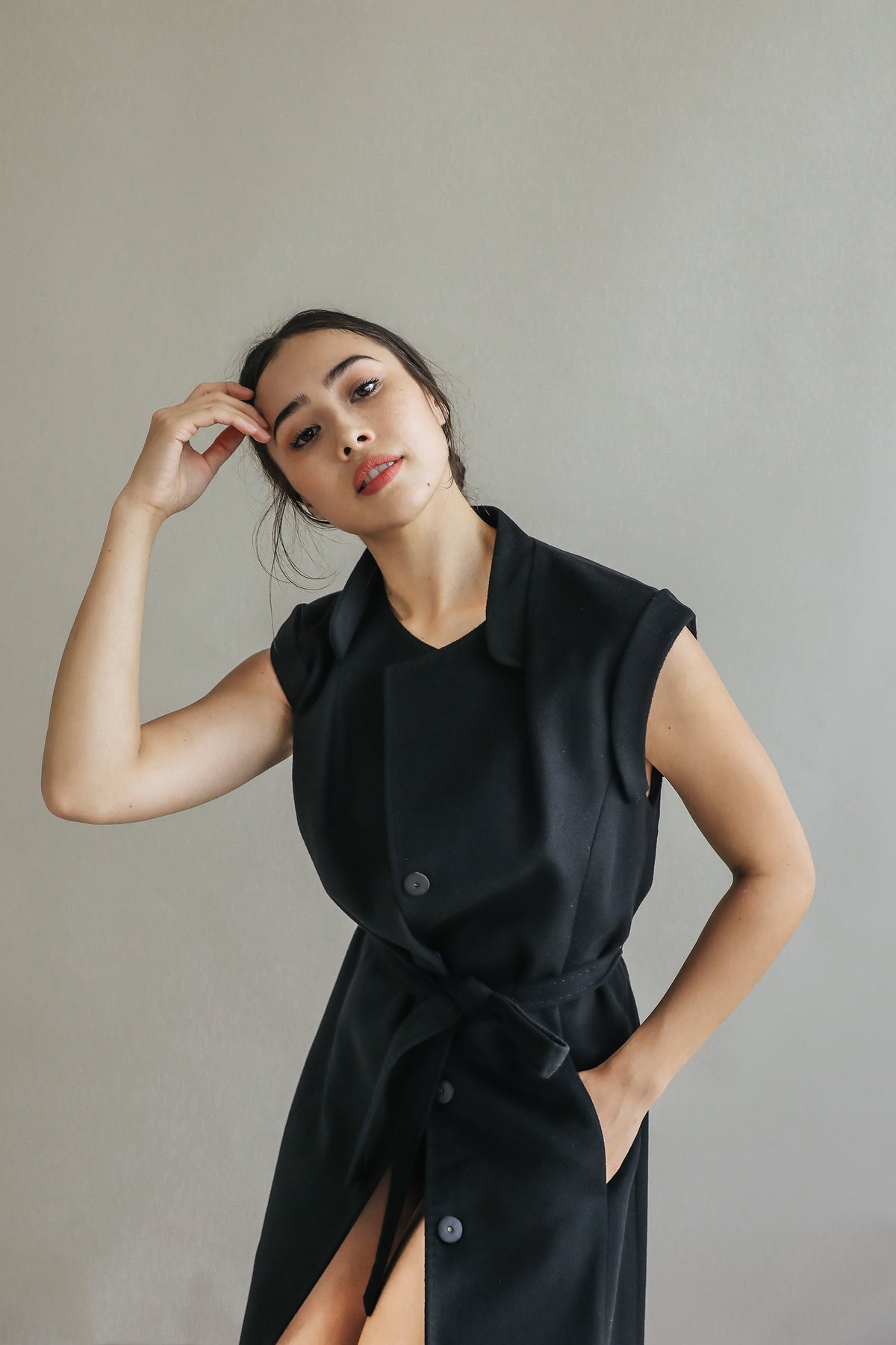 A woman models the Lexington—a sleeveless black dress with a belted waist—posing against a plain light gray background, one hand in her pocket and the other gently touching her forehead.