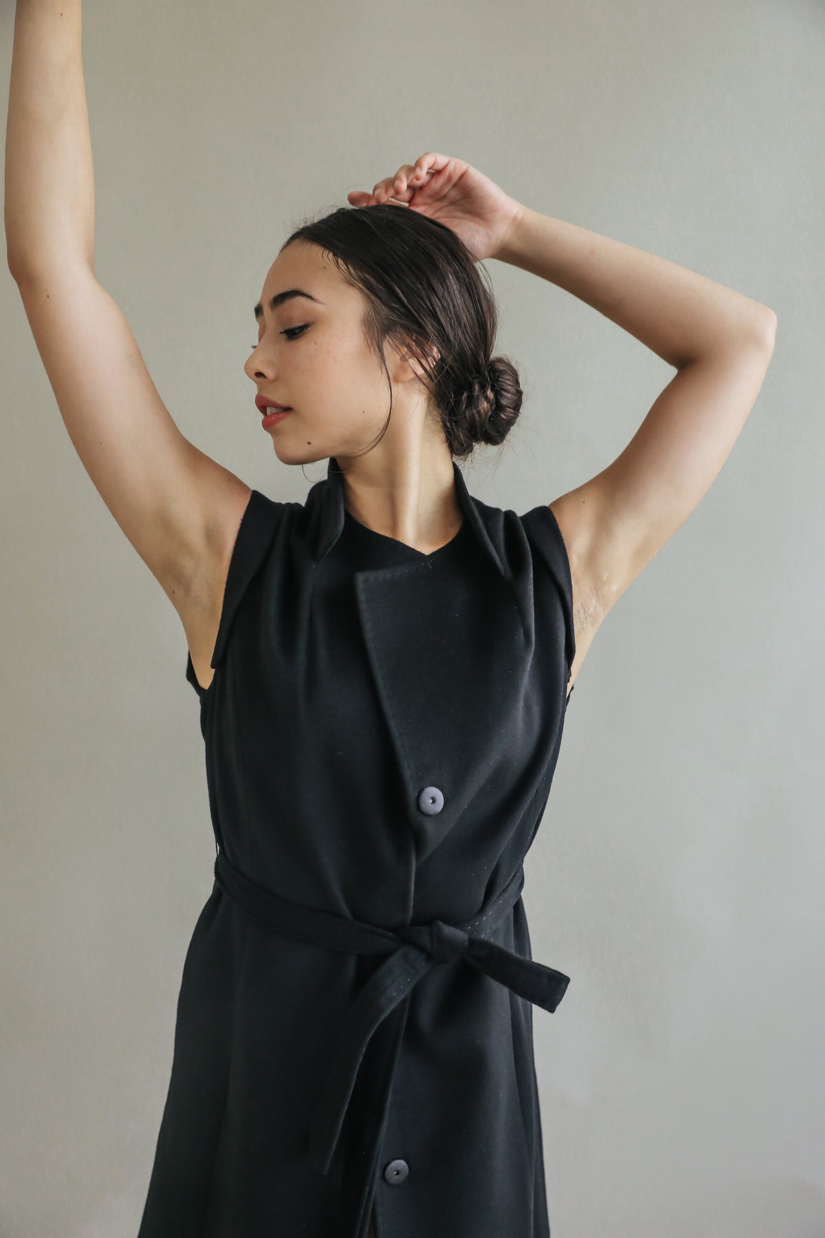 A woman with dark hair in a low bun poses gracefully with her arms raised and crossed, wearing the Lexington sleeveless black dress with a belted waistline against a plain light background.