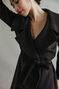 A person with dark hair poses in the Malvern, a luxurious double-breasted coat, with one arm raised. The neutral background and downward gaze create a contemplative atmosphere, highlighting the sophisticated elegance of the woven cashmere fabric.