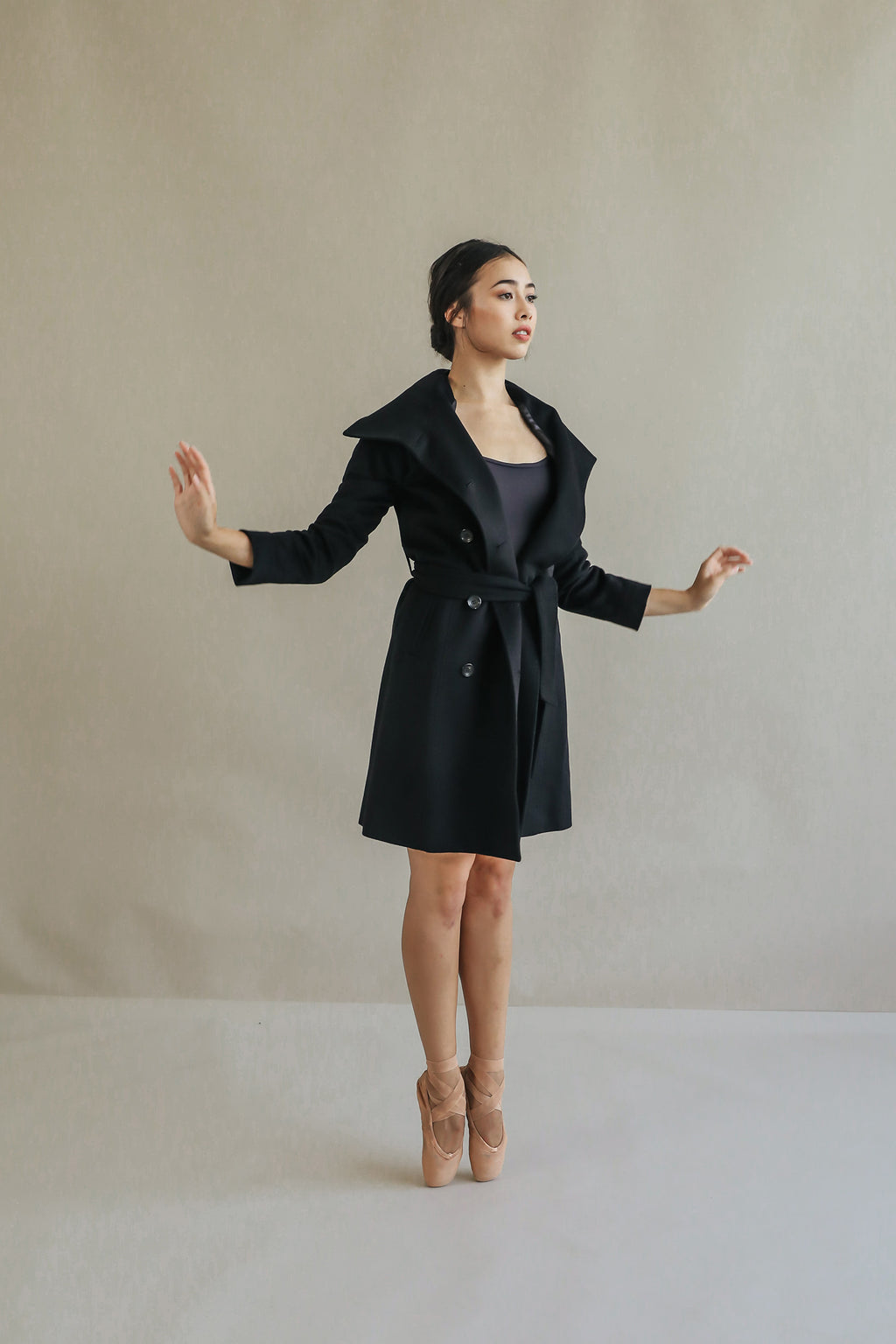 A person stands gracefully on pointe, clad in the Marylebone black cashmere coat over a leotard and ballet shoes, against a plain, neutral-toned wall.