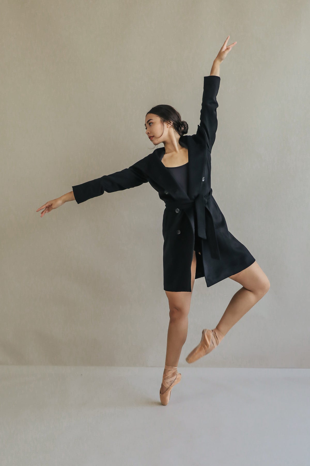A dancer elegantly poses in the Marylebone, a double-breasted trench coat, and ballet shoes against a plain background, balancing on one foot with arms extended gracefully.