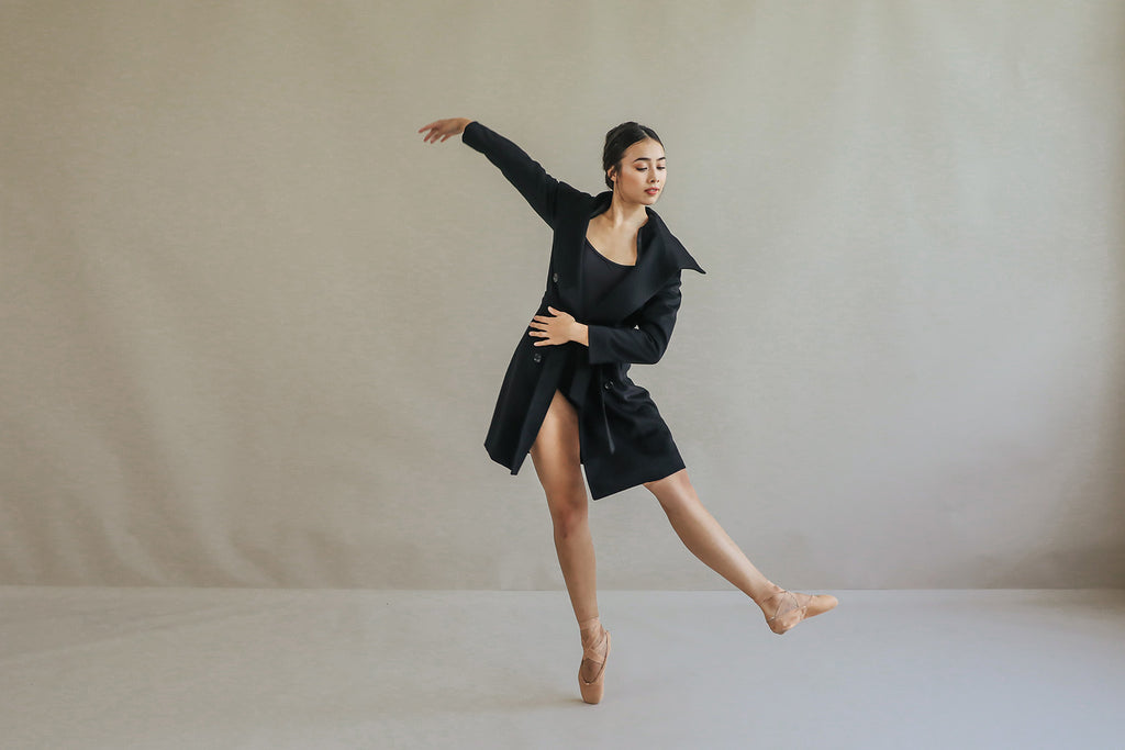 Wearing the elegant Marylebone black cashmere coat, a dancer on pointe shoes performs a ballet pose against a neutral backdrop. One arm extends overhead, while the other rests on her waist, highlighting her balance and grace with an outstretched leg.