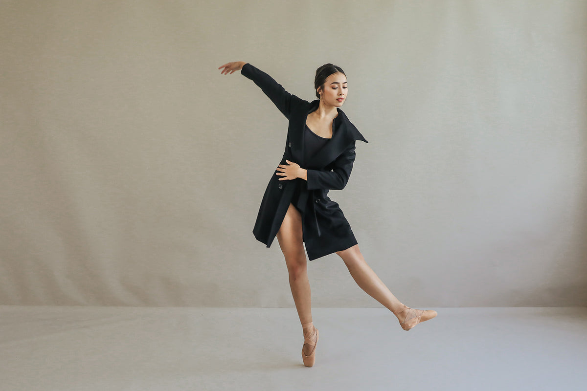 Wearing the elegant Marylebone black cashmere coat, a dancer on pointe shoes performs a ballet pose against a neutral backdrop. One arm extends overhead, while the other rests on her waist, highlighting her balance and grace with an outstretched leg.