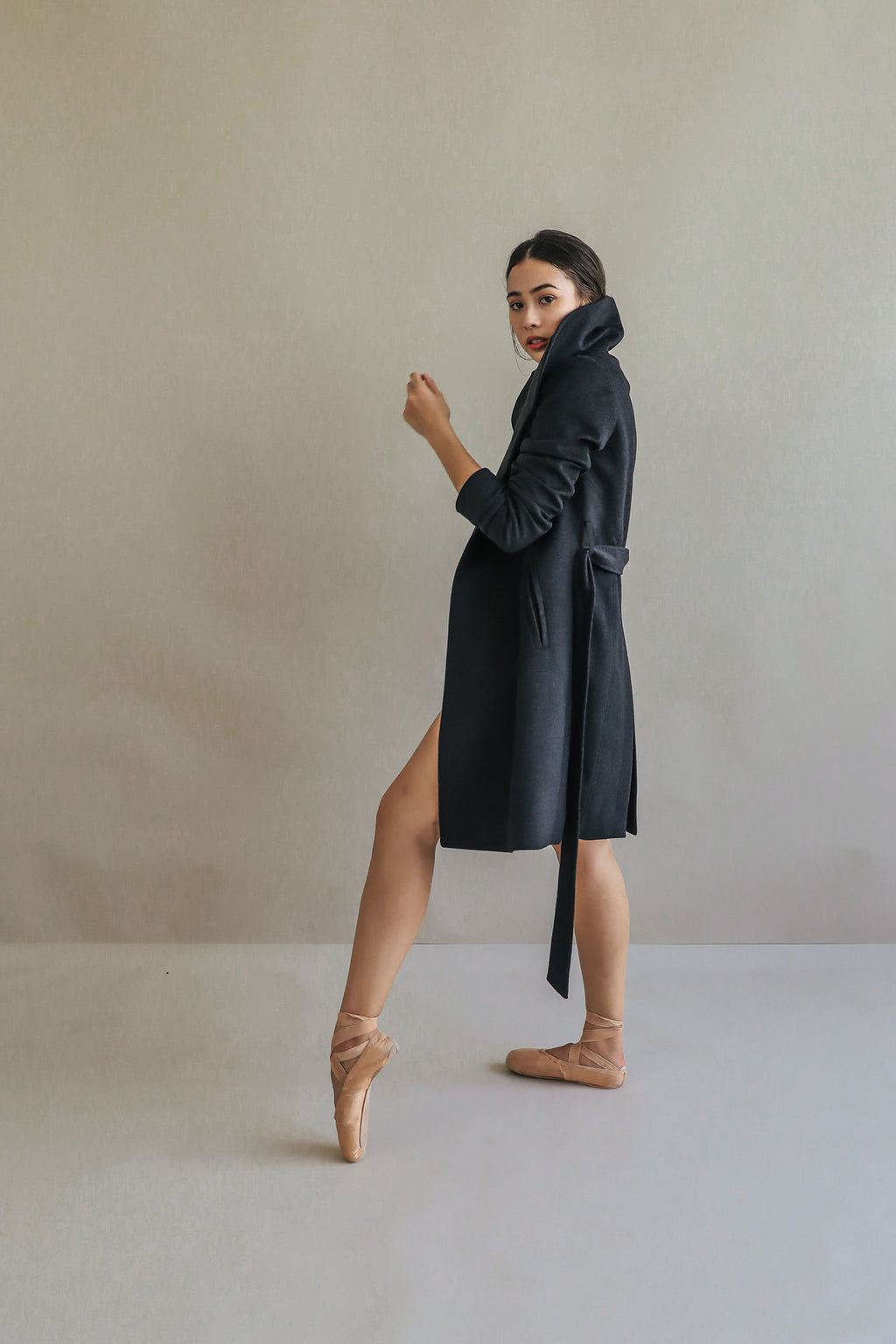 A ballerina in the Marylebone cashmere trench coat poses gracefully on pointe. She stands with one leg extended, looking over her shoulder against a neutral background. Her hair is tied back, and she wears tan ballet shoes.