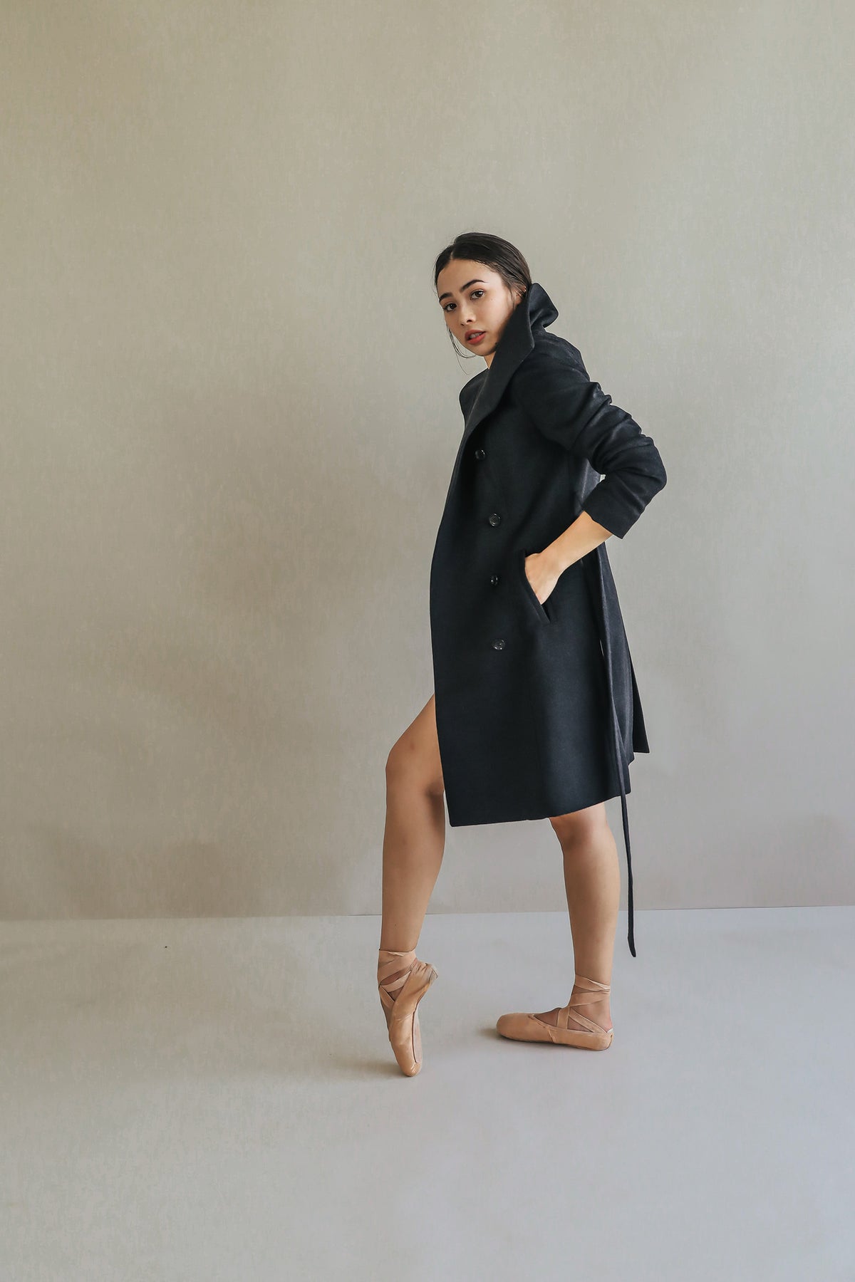 A woman stands en pointe in a Marylebone double-breasted black coat, with her hands in pockets and an intense gaze at the camera, set against a plain neutral background that highlights her poised stance.