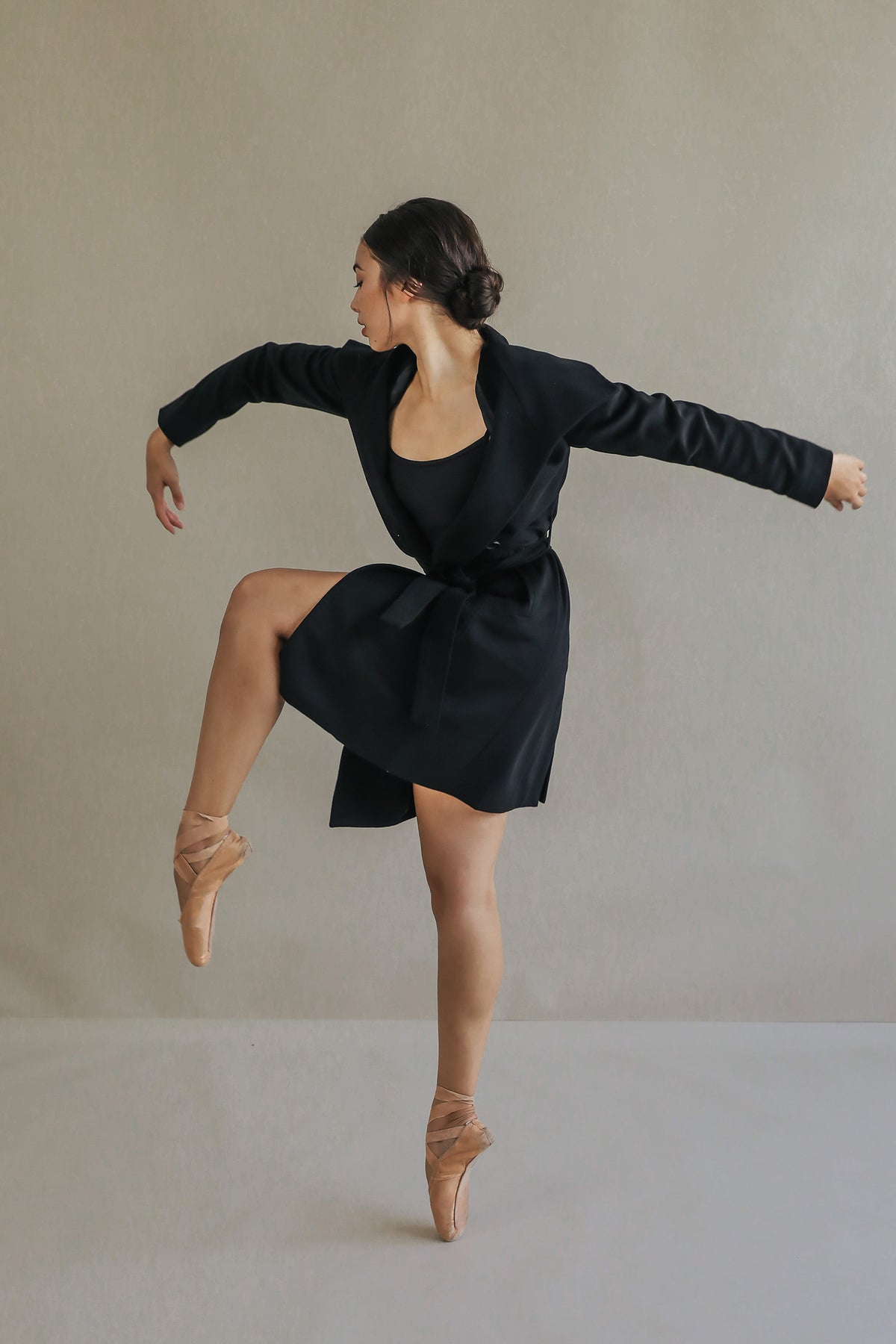 A ballerina in a Marylebone black cashmere outfit stands on pointe, gracefully lifting one leg and extending her arms. The plain, light background accentuates her elegant pose and fluid movement.