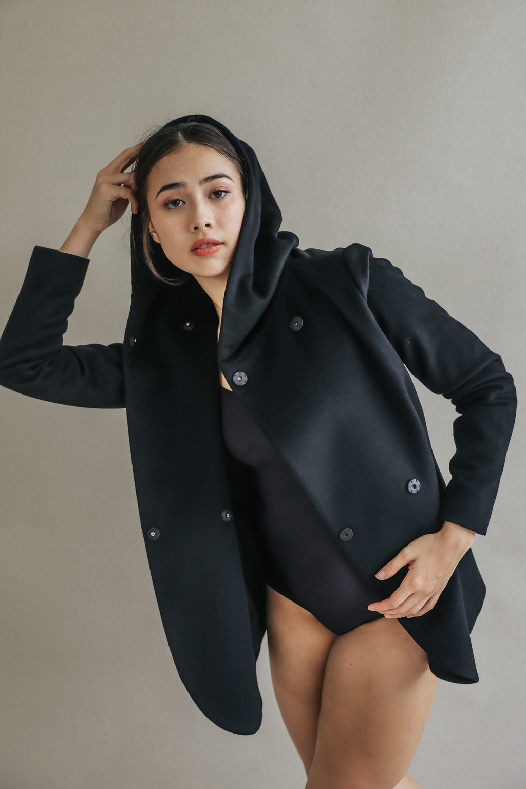 A woman models the Mitchley black cashmere hooded coat over a black bodysuit, posing against a plain light background with one hand on her head and the other on her hip for an effortlessly luxurious look.