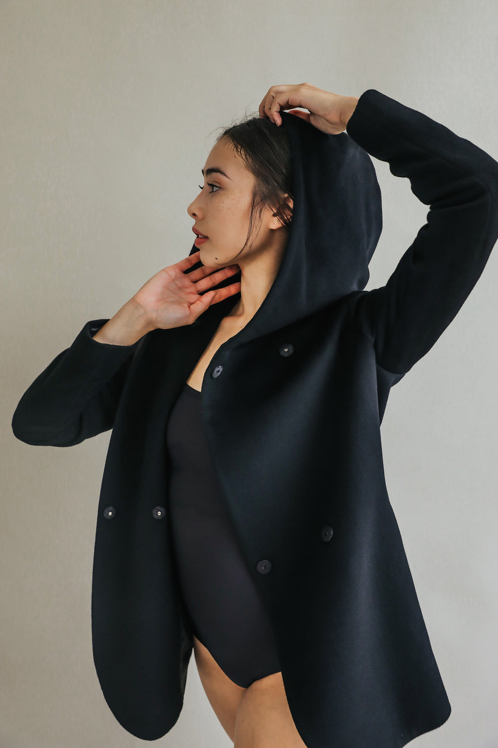 Wearing the Mitchley, a person in black poses with one arm raised to adjust the hood, looking to the side against a plain light background.