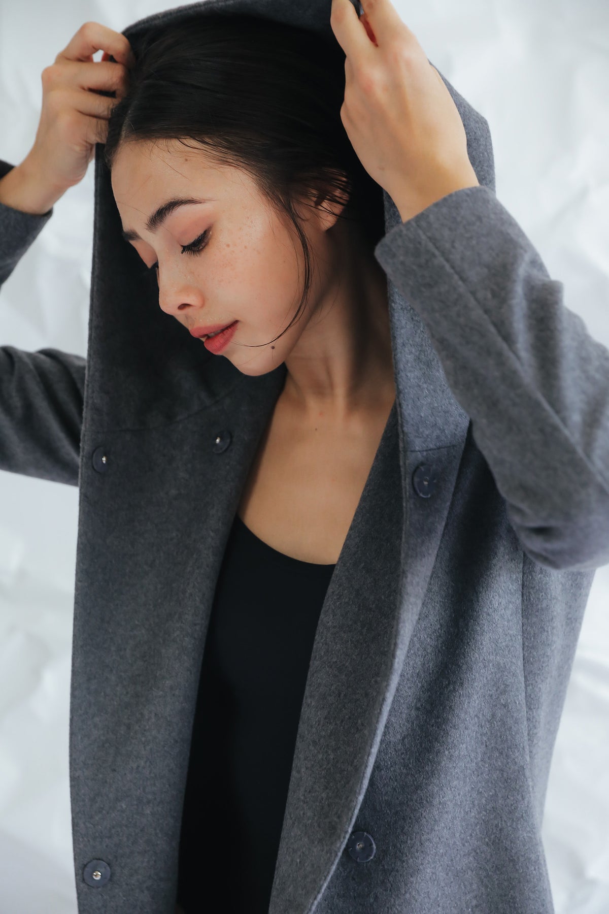 Wearing a Mitchley Long coat, a person adjusts the hood with a serene expression. The gray woven cashmere and handmade snap buttons add charm against the light, textured background.