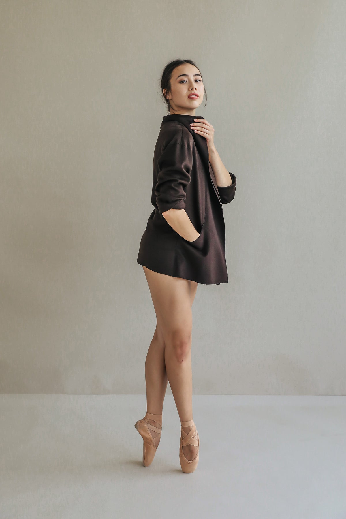 Draped in an oversized Mornington woven cashmere blazer with raw-edge stitches, a woman poses gracefully on pointe in ballet shoes, standing against a neutral background and exuding elegance and balance.