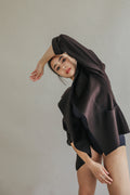 A person poses elegantly against a gray background, wearing the Mornington, a dark, woven cashmere garment featuring raw-edge stitches. Their arms are gracefully positioned, one bent over their head, creating an artistic and dynamic composition.