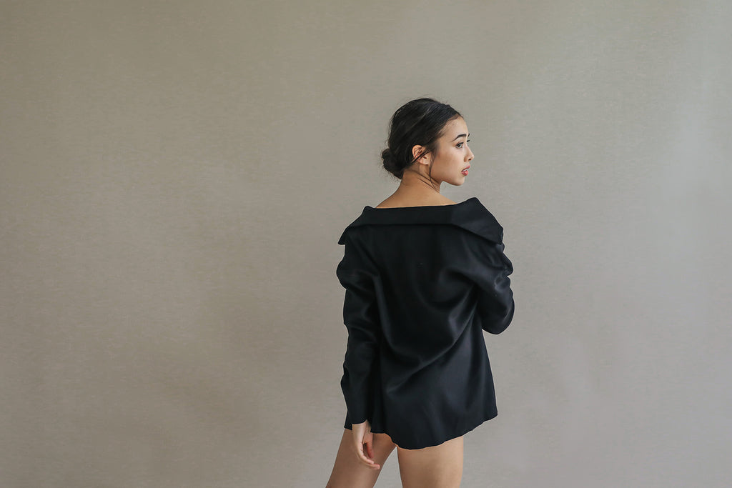 A person with dark hair stands against a neutral background, dressed in the oversized black Mornington shirt featuring raw-edge stitches. The person is slightly turned sideways, glancing over their shoulder.