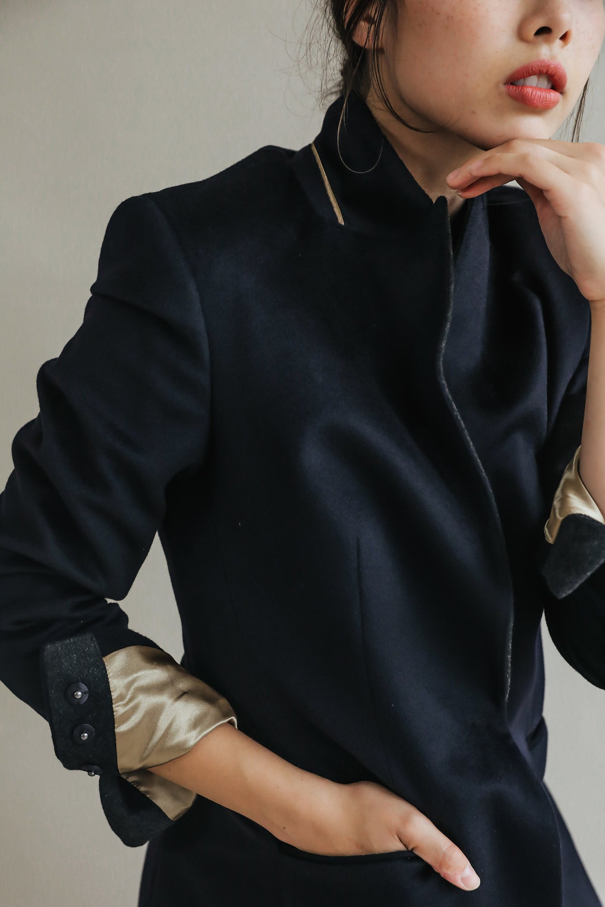 A person stands with one hand in their Sloane coat pocket and the other touching their chin. They are wearing a dark, buttoned Sloane coat with structured shoulders and light-colored lining visible at the sleeves. The background is neutral, emphasizing their upper body and the finely woven cashmere details of the Sloane coat.