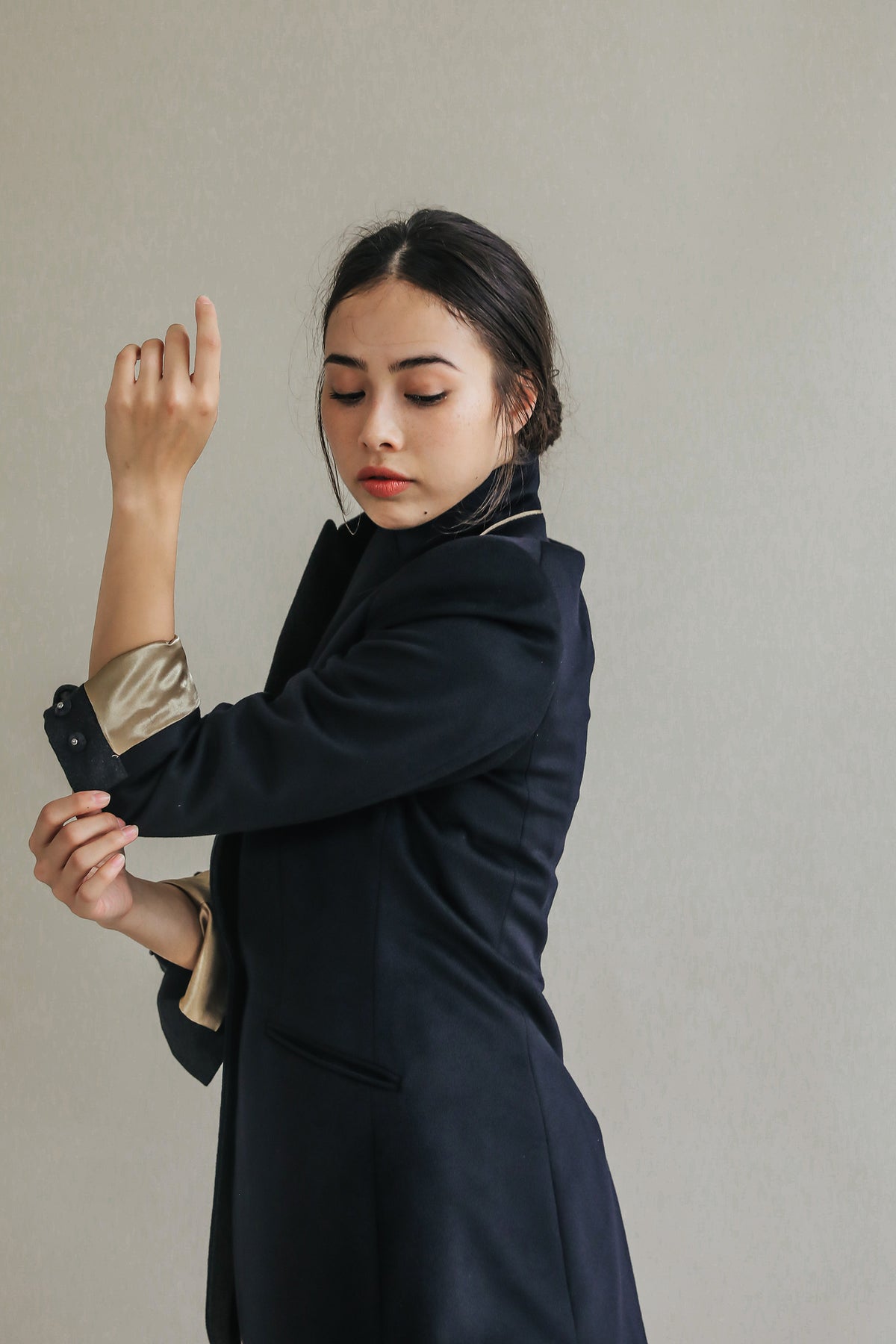 The woman with long dark hair in a low bun is adjusting the sleeve of her Sloane blazer, which features structured shoulders. She has a focused expression and is looking down slightly against a plain, light-colored background.