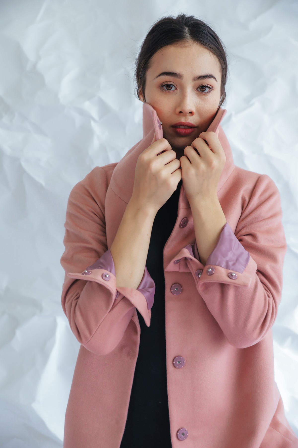 A woman with dark hair stands against a crumpled white background, wearing the Stamford II—a light pink, high-collar coat with buttons. She holds the collar up with both hands. This signature jacket, crafted from woven cashmere, allows her to gaze towards the camera with a neutral expression.