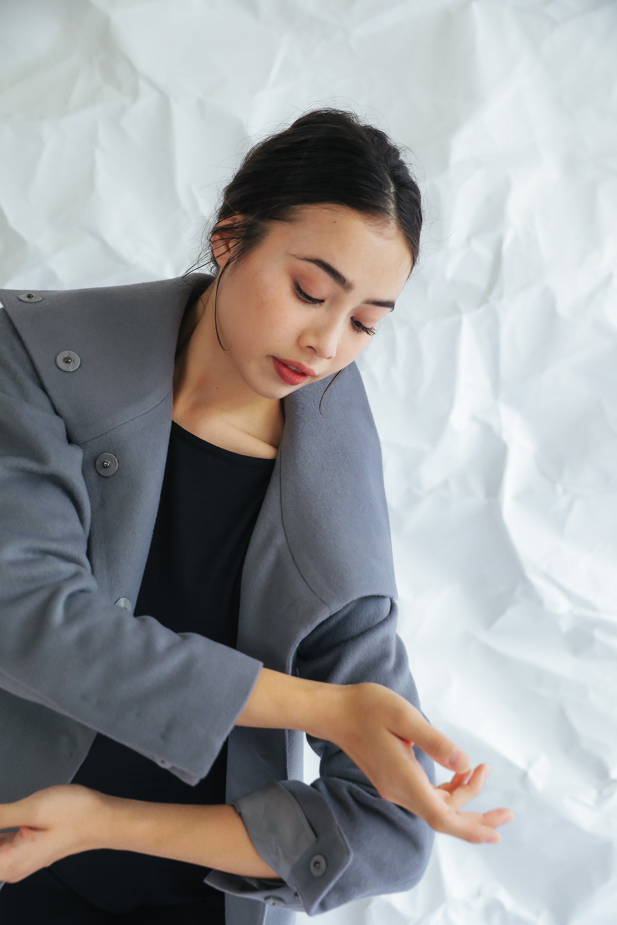 A woman with long dark hair, donning the Stamford II grey high-collar coat over a black top, poses against a white crumpled paper background. She gazes downward with a serene expression, her left hand softly resting on her right arm.