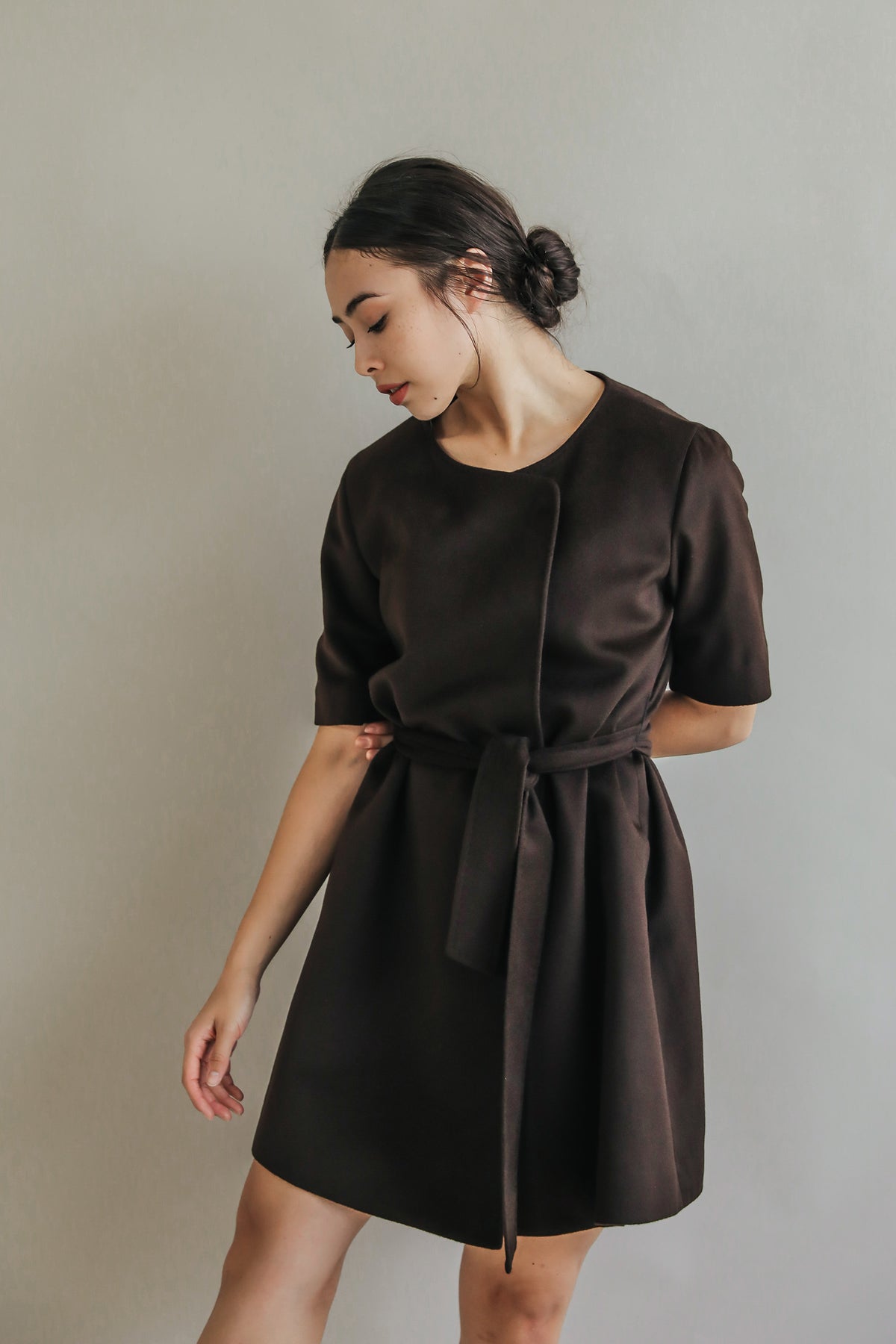 A person elegantly poses against a neutral backdrop, wearing the Wexley dress—a dark brown A-line with short sleeves and a waist tie. Their hair is neatly tied back as they gaze down thoughtfully, one hand gracefully raised in delicate poise.
