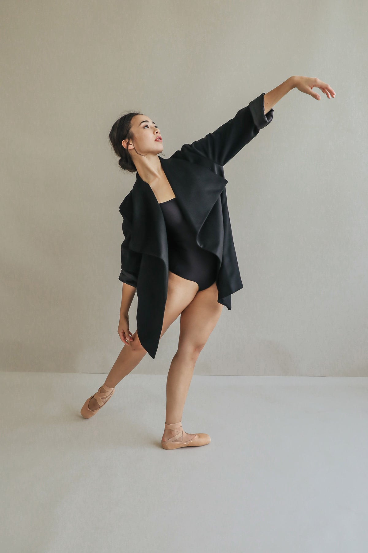 A ballet dancer in the Winston black leotard and a draped-collar, relaxed-fit jacket poses gracefully on pointe with one arm extended upward and the other bent, standing on one leg against a plain light background.
