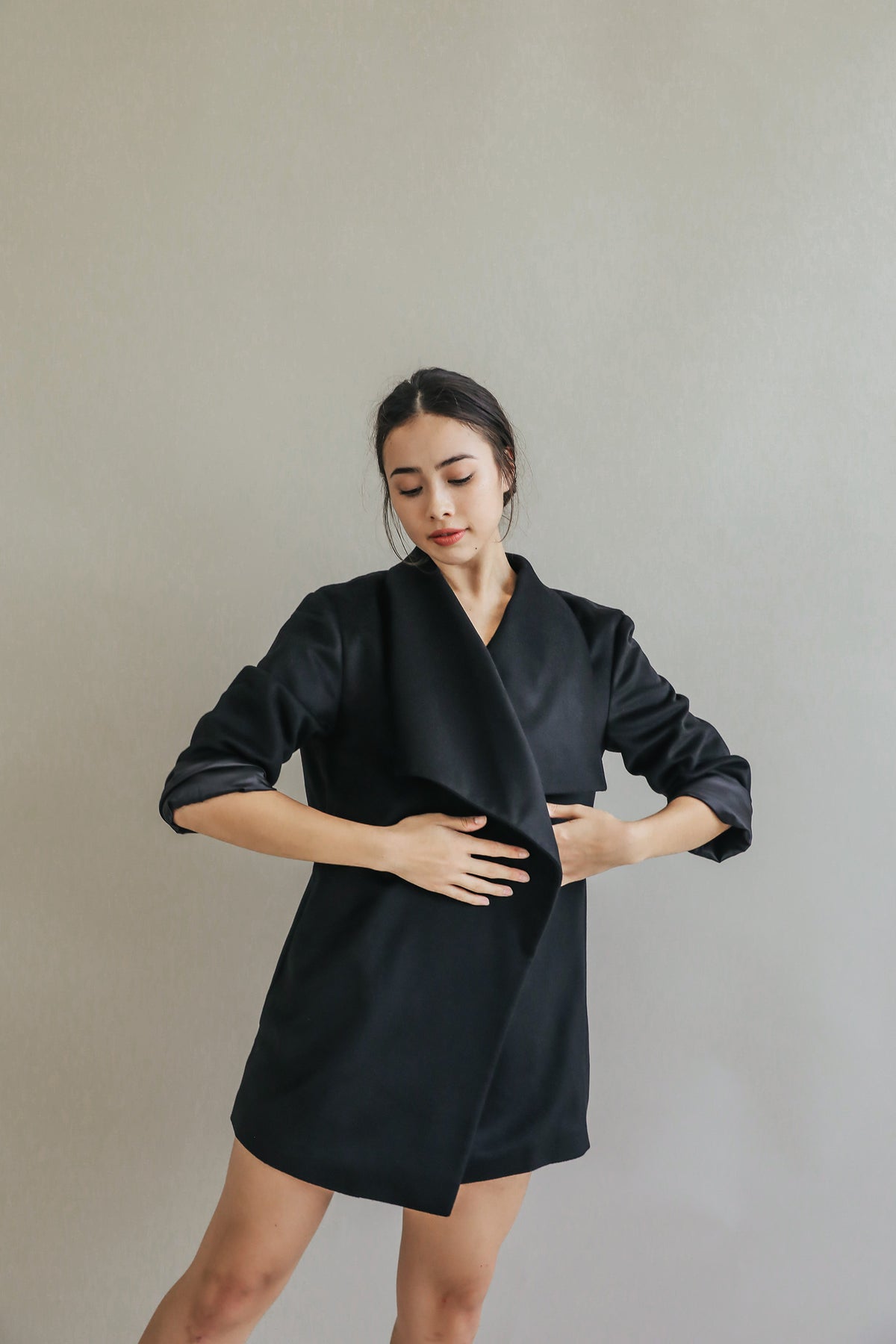 A woman models the Winston dress, a long-sleeved black wrap style with a relaxed fit, standing against a plain light gray background and looking down with one arm bent at her waist and the other resting at her side.