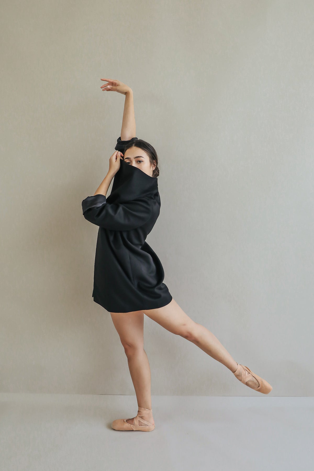 Wearing the relaxed fit Winston black shirt and ballet shoes, a person poses gracefully on one leg with one arm raised and the other partially covering their face against a plain light background.