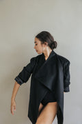 A person with dark hair in a low bun poses gracefully against a plain light background, wearing the Winston relaxed fit black blazer and black bodysuit, looking down and to the side with one arm extended.