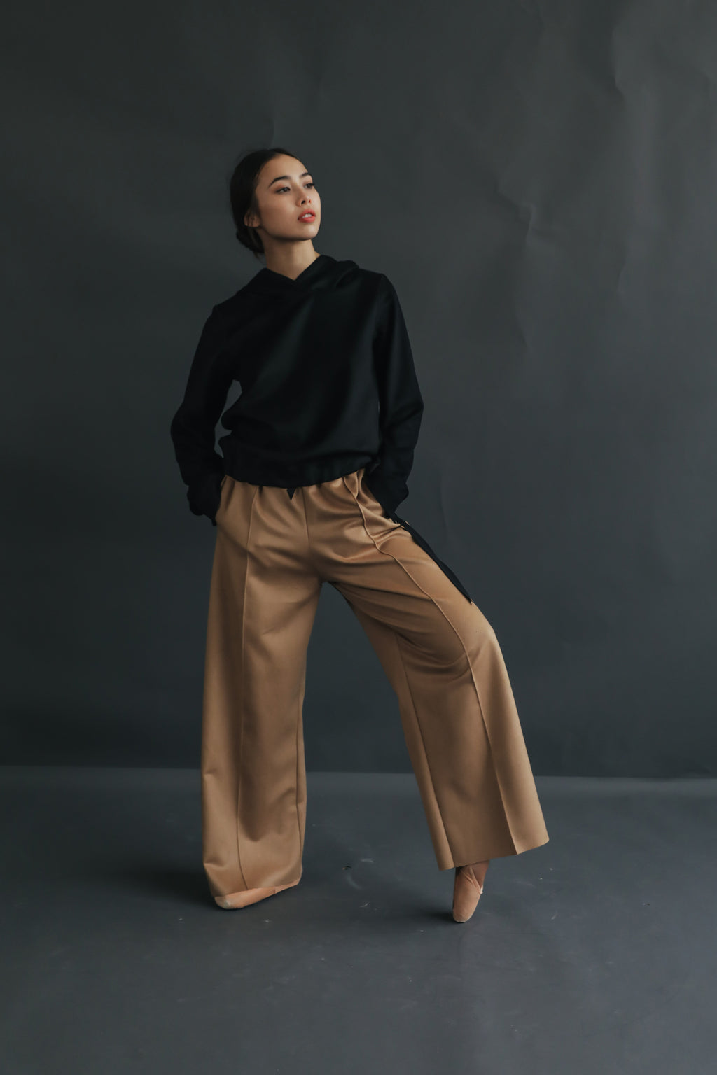 A woman poses confidently against a dark gray backdrop. She is wearing a black long-sleeved top and tan woven Cashmere Trousers. One hand is in her pocket and she is gazing upward with a thoughtful expression.