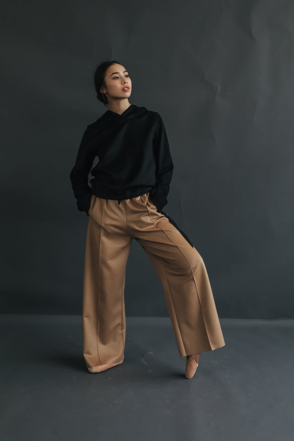 A woman poses confidently against a dark gray backdrop. She is wearing a black long-sleeved top and tan woven Cashmere Trousers. One hand is in her pocket and she is gazing upward with a thoughtful expression.