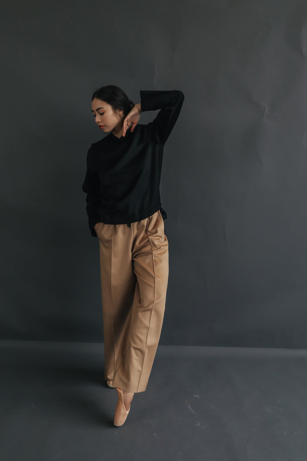 A person stands against a dark grey background, posing with one hand near their neck and the other arm relaxed. They are wearing a black long-sleeve top and loose beige, straight leg Cashmere Trousers, and they have their eyes closed.