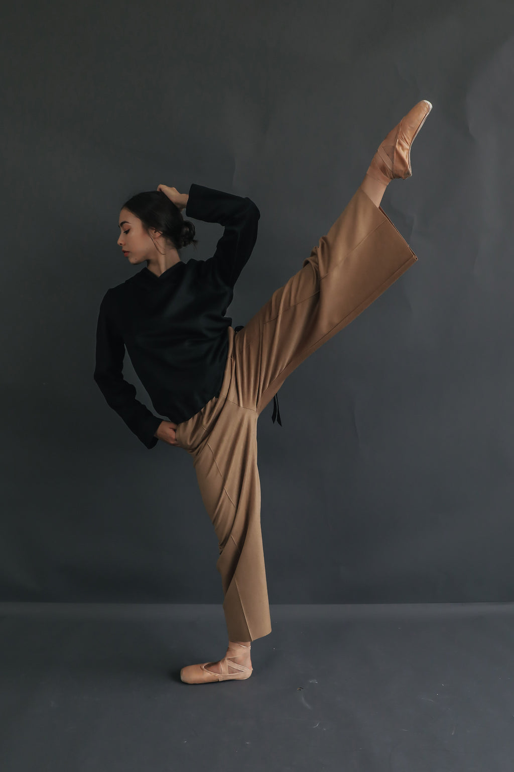 A person dressed in a black long-sleeve top and light brown Cashmere Trousers performs a high kick. They stand on their left leg with their right leg extended straight up and their right hand holding their left ankle, against a plain dark background.