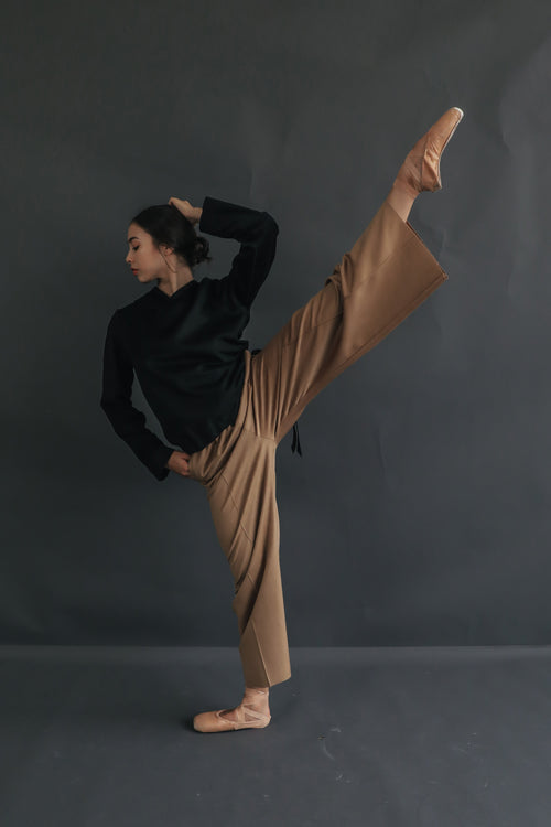 A person dressed in a black long-sleeve top and light brown Cashmere Trousers performs a high kick. They stand on their left leg with their right leg extended straight up and their right hand holding their left ankle, against a plain dark background.