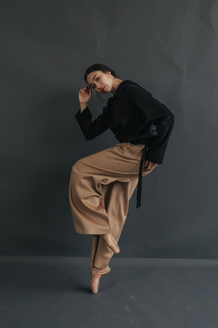 A person poses against a dark grey backdrop wearing a black long-sleeve top, beige Cashmere Trousers, and ballet shoes. They strike a dynamic pose, balancing on one foot and leaning slightly to the side with one arm bent upward and the other resting on their hip.