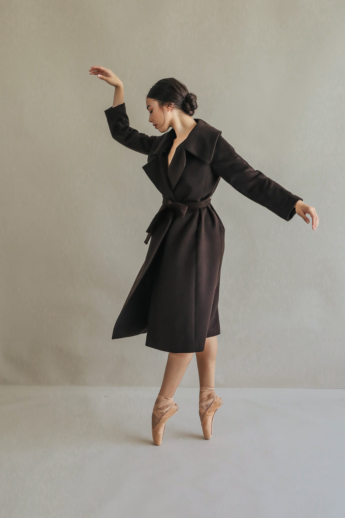 A person gracefully poses on tiptoes in ballet attire, wearing the luxurious Malvern woven cashmere coat. The neutral gray background highlights the elegance and balance of their posture, with arms softly extended.