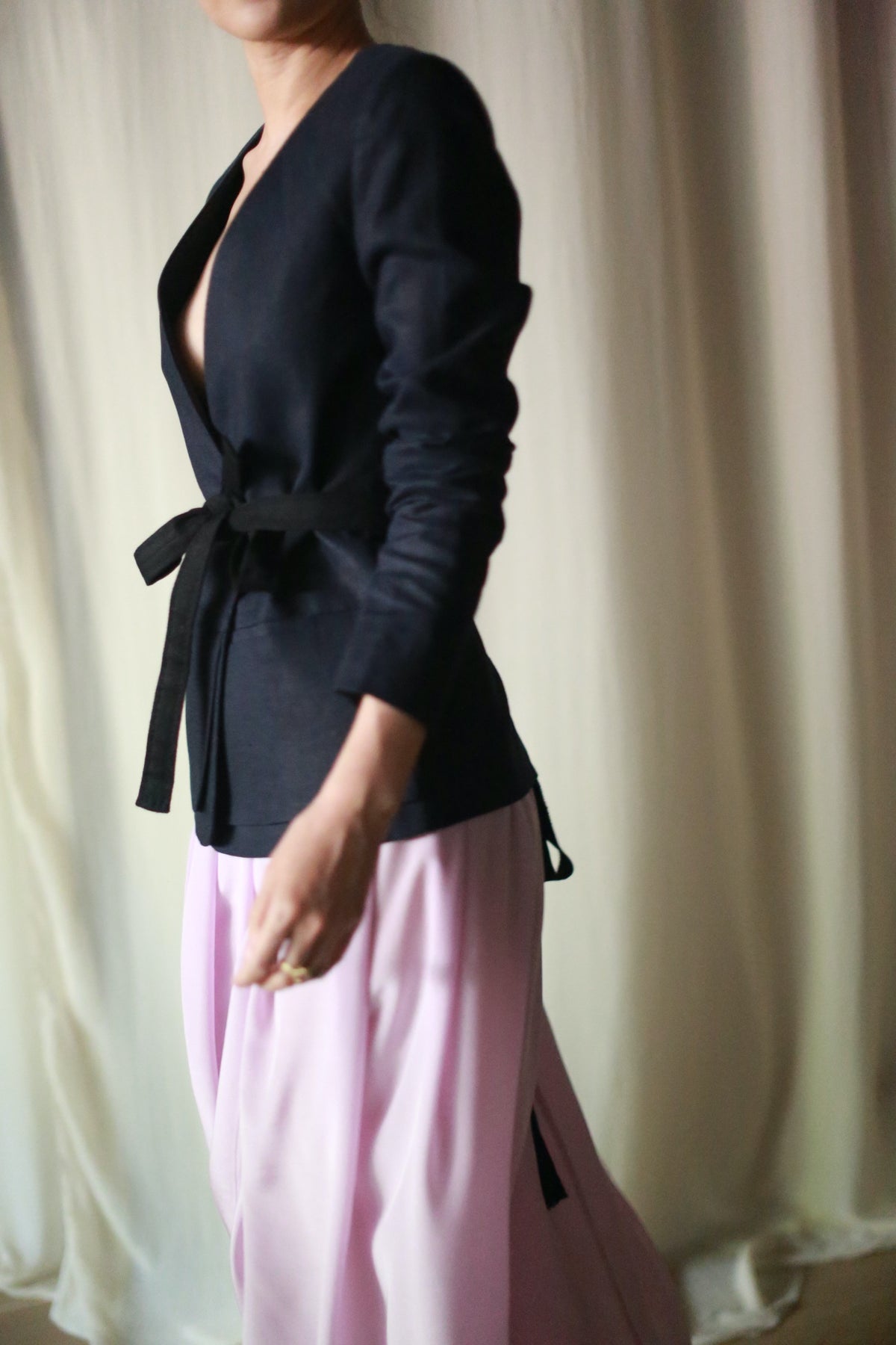 A person stands against a draped white background, wearing a dark Navy Linen Blazer with a tied belt and flowing, light pink pants. The image focuses on their torso and legs, leaving their head out of frame.