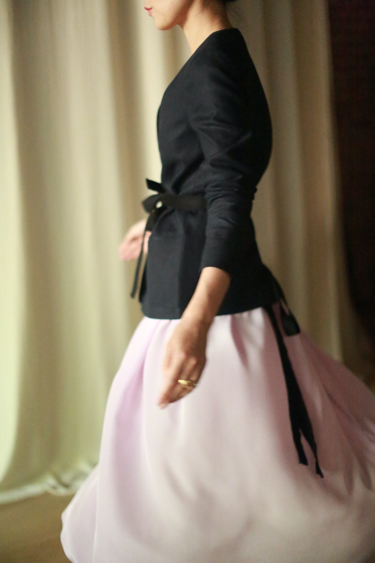 A person in a side profile shot is walking. They are wearing a navy blazer with a black ribbon tied at the waist and a light pink, silk pleated wrap skirt. The background is softly lit with curtains, creating an overall scene with a gentle and elegant ambiance.