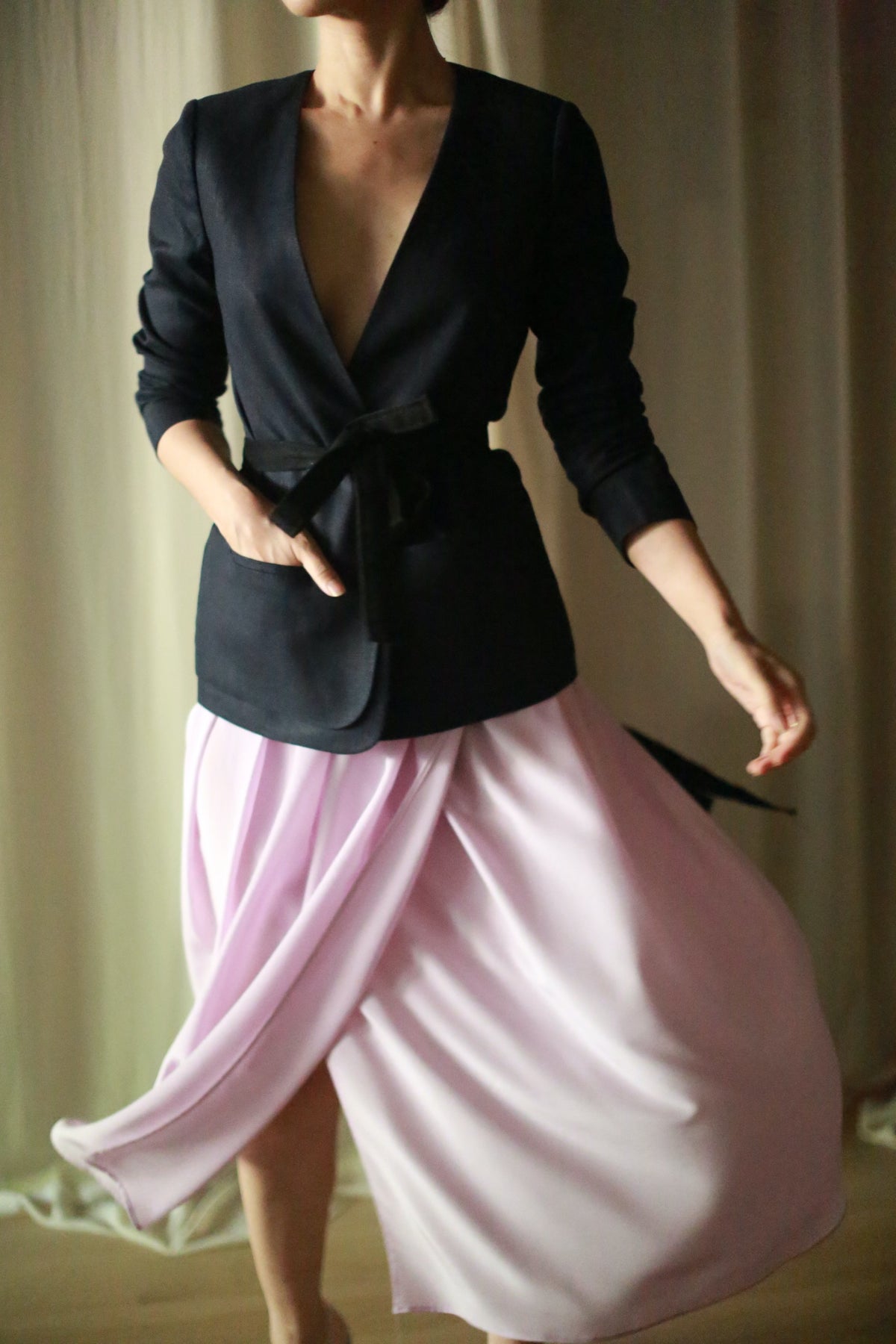 A person is wearing a navy linen blazer and a flowing light pink skirt. The blazer features a deep v-neckline and a belt tied at the waist. The person is standing with one arm gracefully extended, causing the skirt to billow slightly.