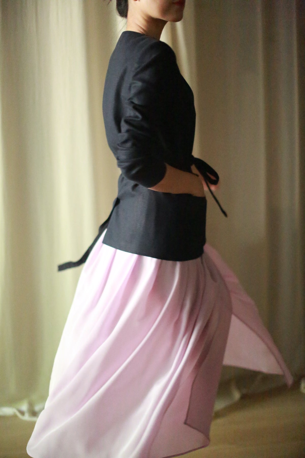A person in a navy linen blazer and flowing silk pleated wrap skirt captured mid-twirl, with motion creating a dynamic blur in the skirt. The background is softly lit, suggesting an indoor setting with curtains. The image conveys a sense of movement and grace.