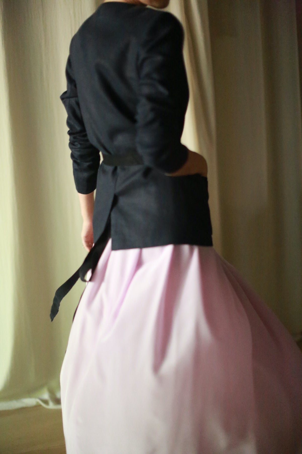 A person is wearing a Navy Linen Blazer and a long, flowing pink skirt. The backdrop is neutral and soft, possibly curtains. The person’s face and upper features are not visible, and they are posing with their back partially turned to the camera.
