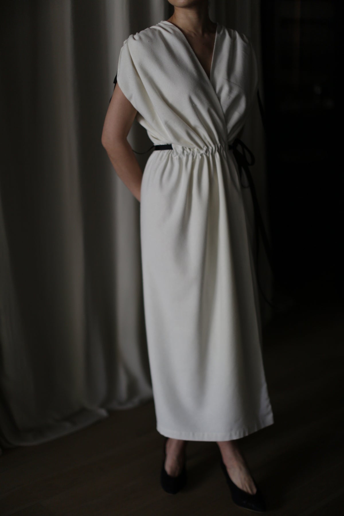 A person wearing the Lune sleeveless handmade white dress with a cinched waist stands with their hands placed behind their back. The figure is partially visible from the neck down to the feet, which are in dark-colored shoes, against a softly lit background.